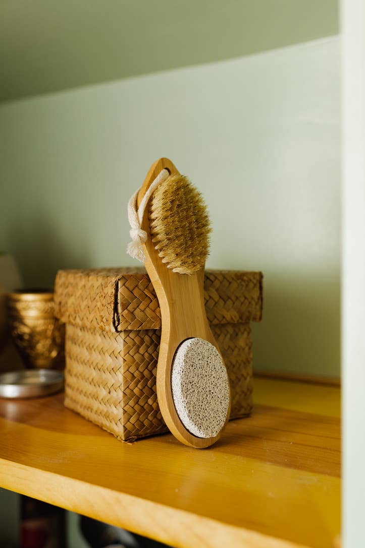 A bamboo exfoliating foot pedicure tool with boar bristles, a pumice stone, and metal filer, displayed on a wooden shelf.