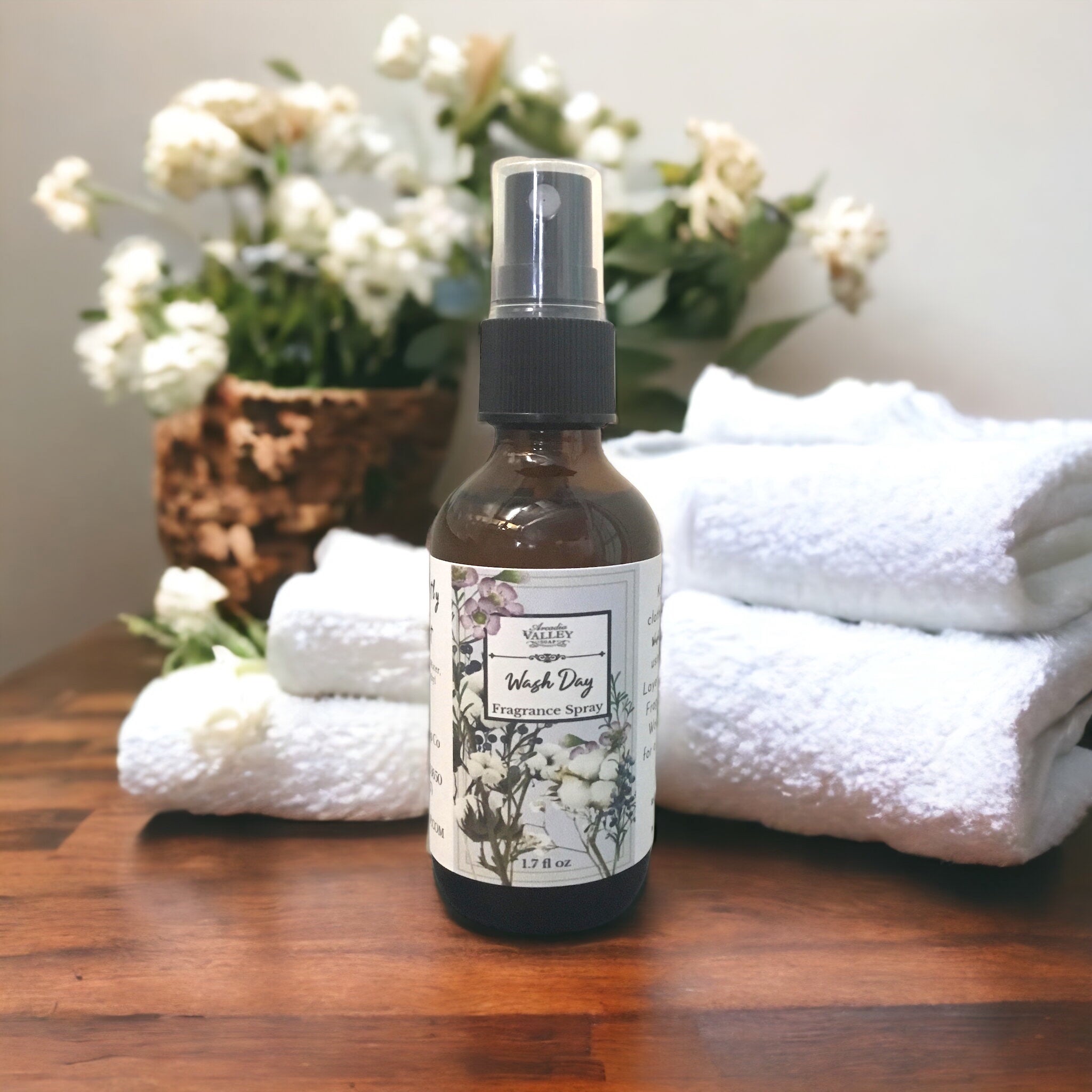 Wash Day Fragrance Spray in a glass amber bottle with plastic mister and floral decorative label on a wooden table with white towels and a basket of flowers in the background